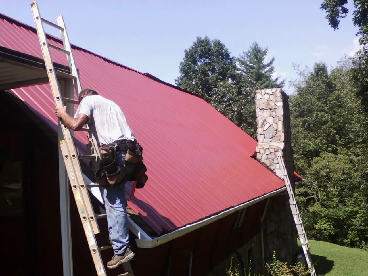 Roofer on a ladder beside a red metal roof and stone chimney in Albany